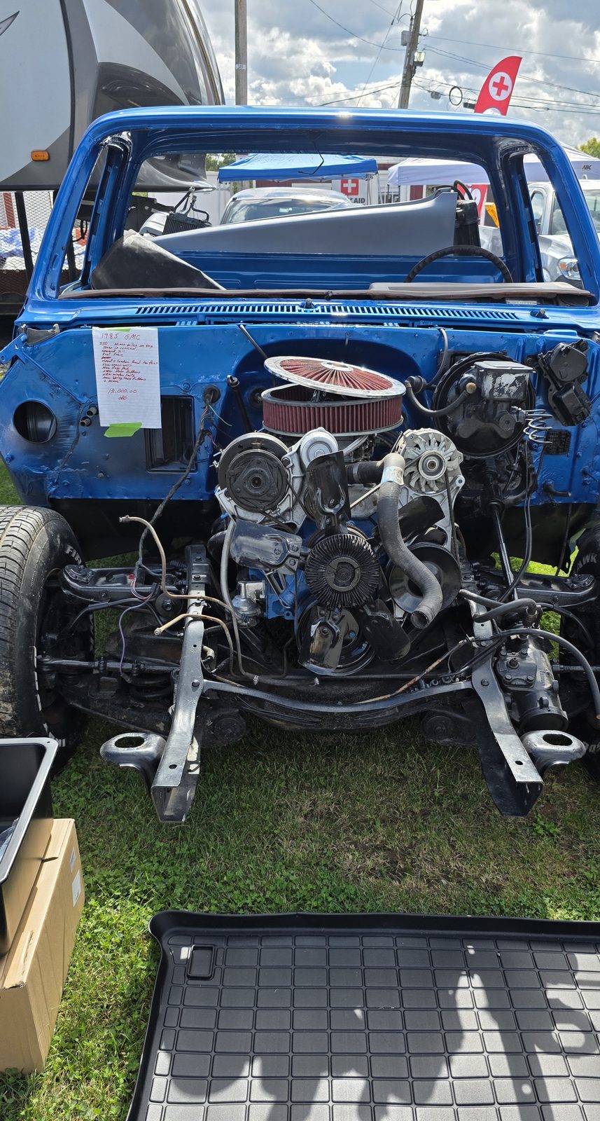 A blue 1985 GMC squarebody cab and frame with the hood, fenders, and grille removed, showing the small-block V8 with a red air cleaner and the bare front suspension, with a handwritten for-sale sheet taped to the cowl